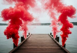 Vibrant crimson smoke clouds drift above a lake, where thick red pyrotechnic plumes erupt from a weathered wooden dock